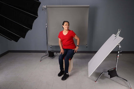 Woman sitting on a stool in a photography studio with C-Stands, backdrop, and lighting equipment.